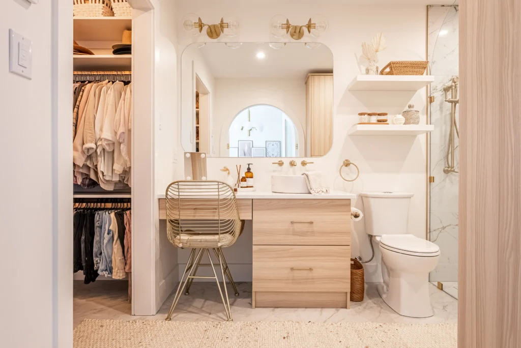 A bright, modern ensuite bathroom connected to an open walk-in closet showcases light wood tones, neutral palette, and boho elements. The closet displays neatly hung pastel clothing on rods, while the bathroom features a vanity with round arch mirror, gold sconces, rattan chair, oak drawers, white sink, toilet, and glass shower. Woven baskets, potted plants, and terrazzo flooring add warmth and texture.
