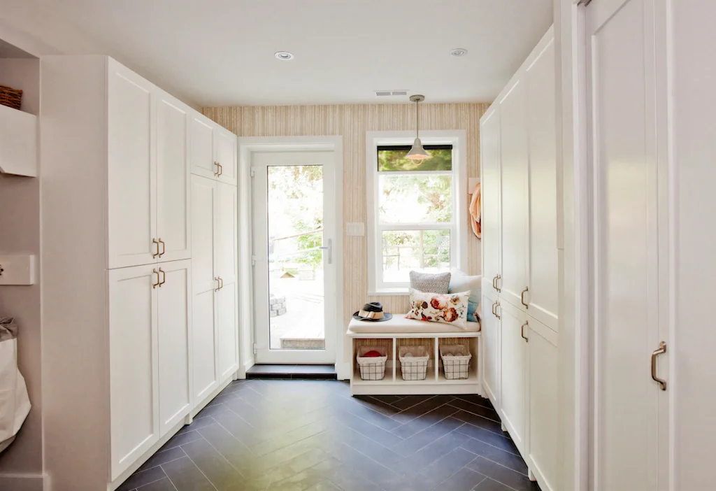 A mudroom with white cabinets, a window, a bench with baskets, and a door leading outside, with dark herringbone flooring
